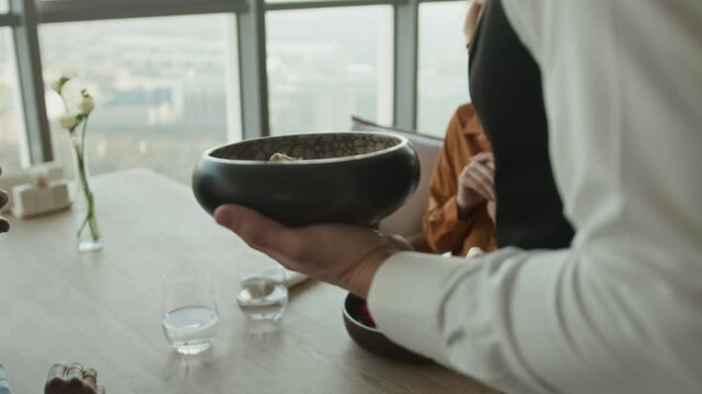 Cropped tracking shot of unrecognizable waiter in classic outfit with bow tie bringing bowls with delicious food and serving female guests sitting at dining table in luxury restaurant