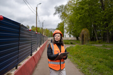 A female civil engineer in an orange construction helmet and vest stands near a fence and writes notes about construction in a notebook