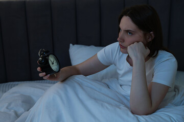 Frustrated young woman with alarm clock suffering from insomnia on bed indoors