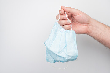 A centred hand, holding a light blue surgical mask by its straps against a plain background...