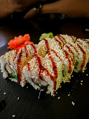Salmon rolls generously sprinkled with sesame seeds and drizzled with teriyaki sauce on a black stone plate, orange pickled ginger and wasabi in the background, Asian food concept with focus on sushi