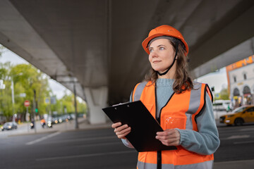 A female civil engineer in an orange construction helmet and vest stands under an overpass against the background of the city with a tablet for notes.Large portrait