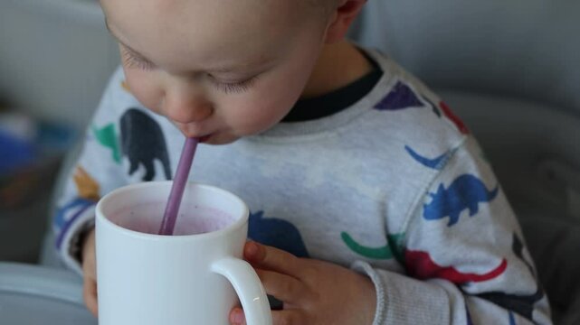 Little boy sitting in high chair and drinking milk shake from a cup through a straw. Closeup, selective focus.
