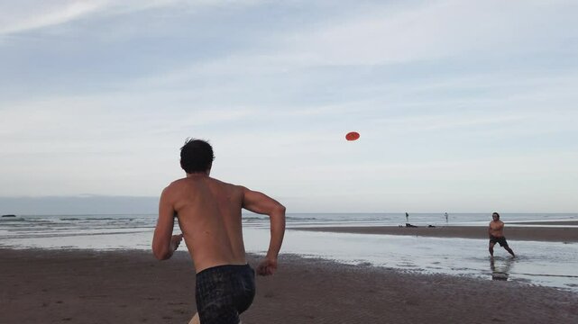 Back view of a shirtless man running toward the sea to catch an airborne orange frisbee as his friend looks on from afar, in Omaha Beach at low tide in dramatic slow motion