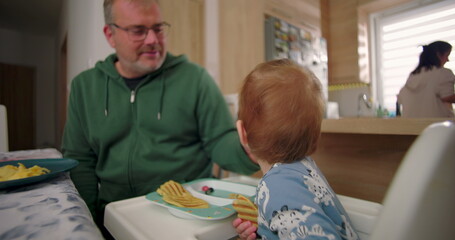 Father watching toddler enjoy breakfast with waffles and fruit, sharing a warm and intimate moment...