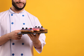 Happy confectioner in uniform holding eclairs on orange background, closeup. Space for text