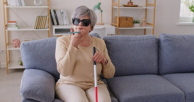 Senior elderly blind woman in black eyeglasses holding cane and using digital assistant on mobile phone sitting on sofa at home. Visually impaired female person talking by mobile phone. 4k video.