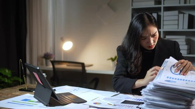 A woman is sitting at a desk with a pile of papers in front of her. She is looking at the papers and she is focused on her work. Concept of productivity and concentration