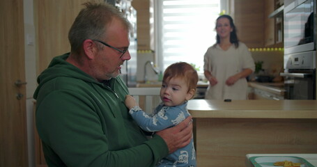 Mother picking up baby from father’s lap, joyful family interaction during a cozy household...
