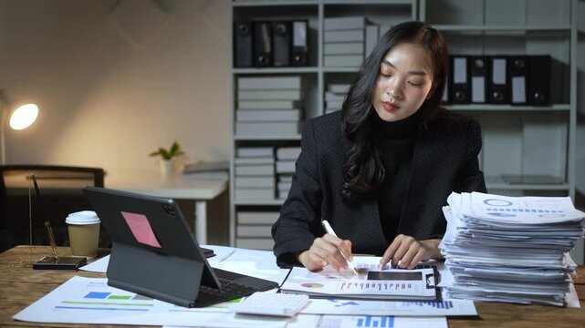 A woman is sitting at a desk with a laptop and a stack of papers. She is writing on a piece of paper with a pen. The scene suggests that she is working on a project or a report