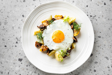 Tasty breakfast with a sunny-side-up egg on roasted vegetables, including mushrooms, potatoes, and corn, garnished with fresh greens, served on a white plate with a textured background.