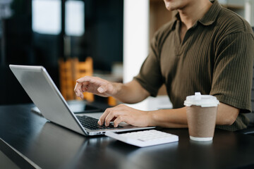 Young man typing on laptop and smartphone at modern office desk with coffee and tablet nearby. Ideal for tech, business