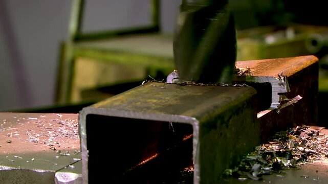 Industrial metal drilling: drill bit cutting through steel profile tube with metal shavings around &mdash; precision machining in workshop