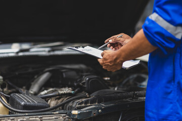 Car mechanic inspecting engine and taking notes on clipboard. Mechanic in a uniform is holding a clipboard and inspecting a car engine, taking notes while assessing the vehicle's condition.