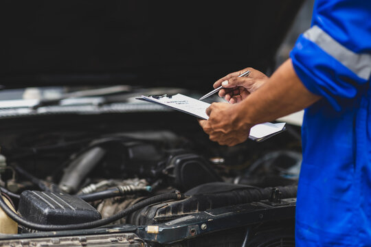 Car mechanic inspecting engine and taking notes on clipboard. Mechanic in a uniform is holding a clipboard and inspecting a car engine, taking notes while assessing the vehicle's condition.