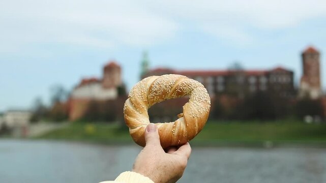 Hand holding a traditional Krakow bagel (obwarzanek) against the backdrop of the historic Wawel Castle and Vistula River. A perfect combination of local food, culture, and travel in Poland.