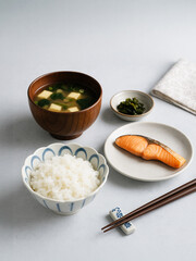 Fototapeta premium Traditional Japanese breakfast with grilled salmon, white rice, miso soup with tofu, and pickled vegetables, served with chopsticks on a light background.