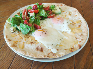 Breakfast plate featuring poached eggs on a flatbread with fresh salad served on a rustic wooden table