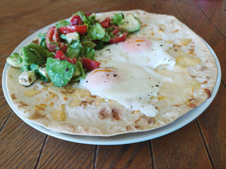 Breakfast plate featuring poached eggs on a flatbread with fresh salad served on a rustic wooden table