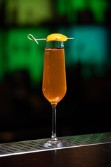 A vibrant orange cocktail is served in a tall, slender glass, garnished with a thin slice of orange peel skewered across the rim.