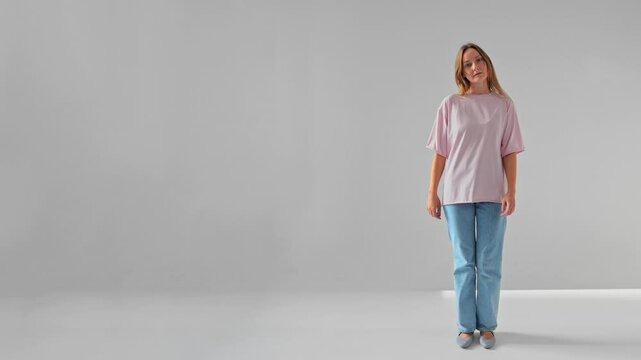 Woman is standing on spot and marching looking at camera filmed in slow motion against white background. Young adult Caucasian lady in blue jeans and pink T-shirt is moving and looking at camera