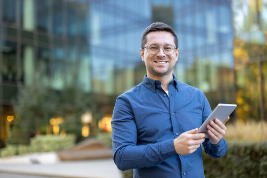 A smiling man, wearing glasses and a blue shirt, holds a tablet in front of a modern building.