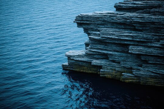 Rocky outcrop meets deep blue ocean. Layered gray stone juts into calm water