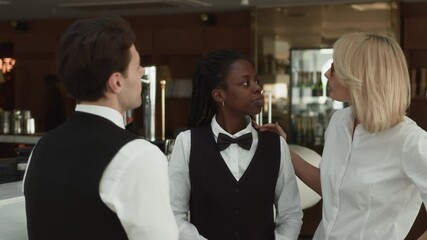 Medium shot of young diverse restaurant staff in elegant classic outfits with bow ties chatting near bar counter, with middle-aged Caucasian female manager joining them discussing working details
