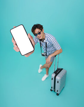 Top view Happy smiling young asian man tourist in summer clothes and baggage showing mockup mobile cell phone app booking hotel isolated on blue background.