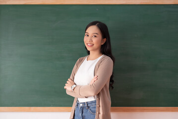 Confident Asian female teacher stands with arms crossed, smiling in a classroom setting beside a...