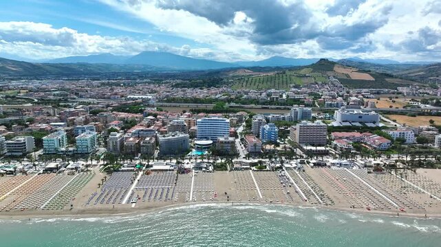 Drone flyover of San Benedetto del Tronto, Italy, showcasing the city, the beautiful Adriatic Sea, and a beach dotted with countless empty sunbeds under the bright sun.