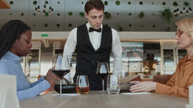 Side view shot of young Caucasian waiter in classic outfit listening respectfully to middle-aged female guest complaining about ordered dish while having lunch with her Black girlfriend in restaurant