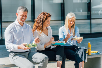 Professional team sharing lunch break outdoors, sitting together near office building, eating from...