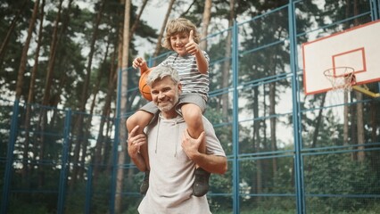 Obraz premium ?ute little boy is sitting on dad's shoulders and holding basketball. Loving father and son smiling, posing and looking at camera on background of basketball court. Family spending time together.