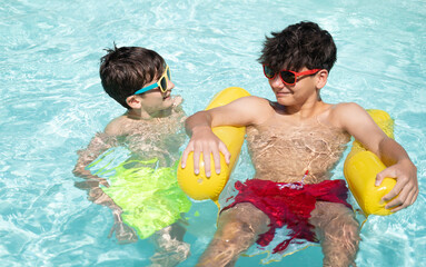 Two brothers wearing sunglasses are enjoying their summer holidays, playing in a swimming pool with an inflatable pool mattress