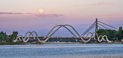 A glowing full moon ascends behind Kyiv’s elegant pedestrian bridge, casting golden reflections across the calm Dnipro River in a serene embrace of night and light.