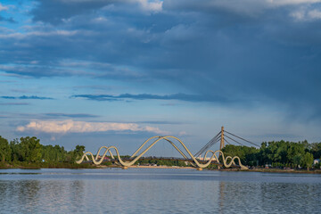 Dramatic clouds hover over the pedestrian bridge on the Dnipro River in Kyiv at sunset. Tranquil cityscape with calm water and soft golden light.