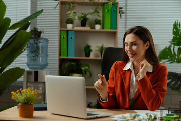Older adult businesswoman works at green office with plants, woman sitting at desk working on laptop, talking online by video call.