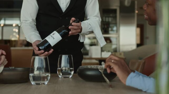 Unrecognizable waiter in classic uniform presenting bottle of wine in luxury restaurant to young African American woman and her guest sitting at dining table enjoying delicious food