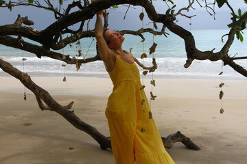 An adult beautiful blonde woman in a yellow long dress walks on the shore of the Andaman Sea and poses under a tree decorated with seashells.