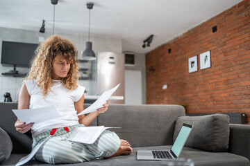 Woman managing home finances sitting on sofa with laptop and documents
