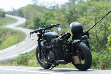 A black motorcycle with travel gear is parked by a winding road through lush, green countryside on a sunny day.