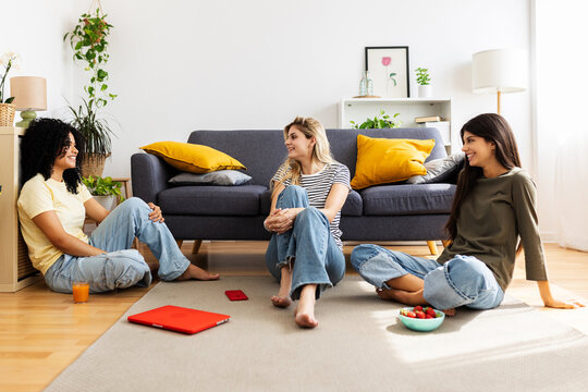 Three happy women are sitting on the floor in the living room, sharing stories and having fun, with snacks and technology nearby. Female friendship and youth concept - Powered by Adobe