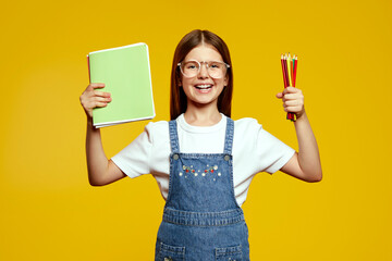 Smart and handsome schoolgirl in trendy denim overalls and trendy glasses holding colored pencils and notebooks. Her joyful smile and bright background radiate learning enthusiasm