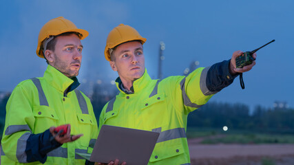 caucasian white engineers in safety uniforms and helmets using laptop and radio for site communication. Concept of teamwork, inspection, and operations at an industrial facility.Construction of Oil, 