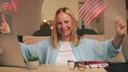 Enthusiastic woman waving two small american flags while sitting at table with laptop, glasses and notebook, expressing patriotism and excitement.USA celebrate 4th of July. Independence Day concept - Powered by Adobe