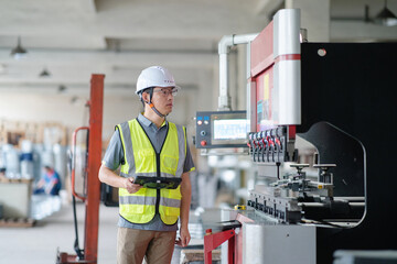 An Asian male engineer is holding a tablet in front of a bending machine.