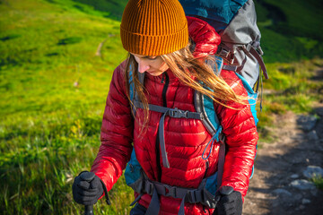 Woman hiking uphill with trekking poles in the mountains, wearing a red jacket and orange beanie,...