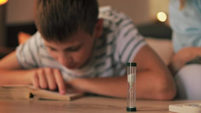 Mother monitoring son's study session, using hourglass timer for creating structured learning environment, promoting concentration and time management skills during quiet evening at home