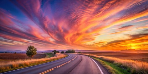Naklejka premium Serene Asphalt Road Winding Through Rural Landscape Under a Vibrant Sunset Sky
